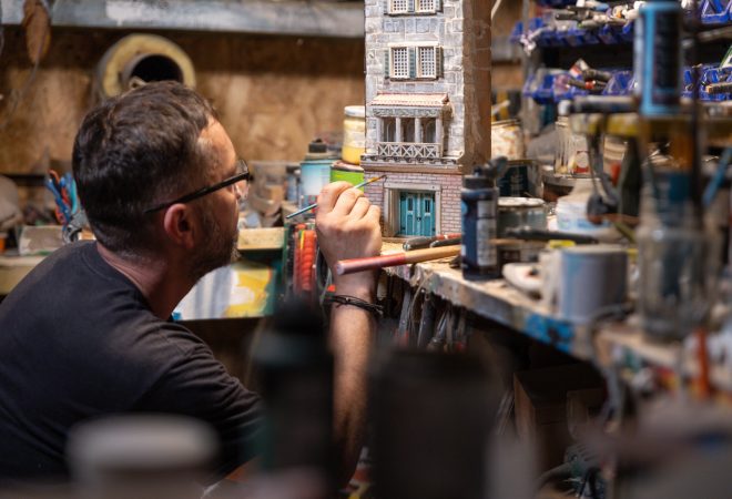 Modeller man working at scale model of miniature building in his workshop full of tools