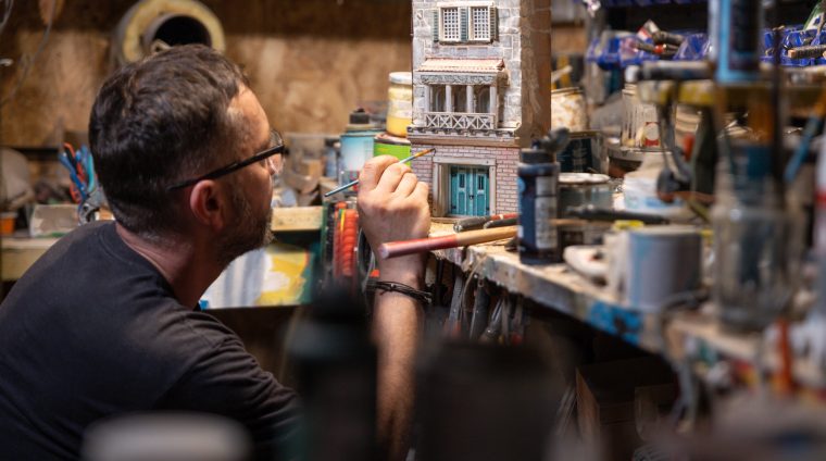 Modeller man working at scale model of miniature building in his workshop full of tools Modeller man working at scale model of miniature building in his workshop full of tools