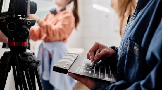 Close-up of hands holding clapperboard while filming with professional camera in indoor setting. Background showing blurred figure working with equipment